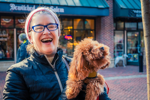A cheerful woman holds a small, curly-haired, brown poodle on a busy street